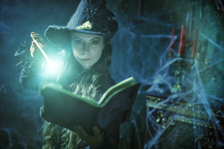 Art of witchcraft. Beautiful curly-haired sorceress in a black hat makes a spell with a magical book and a wand in a dark old castle covered in cobweb. Magic Halloween spells.の写真素材