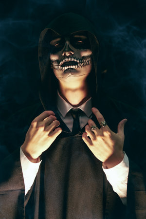 Dangerous man with skull makeup wearing a black robe with a hood stands against a dark mysterious background. Halloween. The devil, death.の写真素材