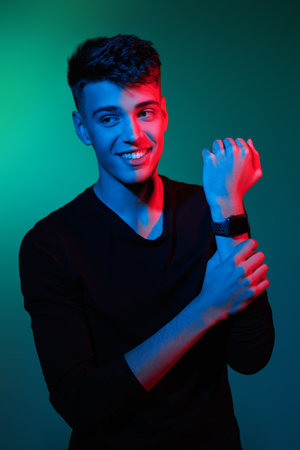 A handsome brunet guy in casual clothes with a watch on his hand looks to the side and smiles joyfully. Green background. Young people, lifestyle. Male beauty.の写真素材