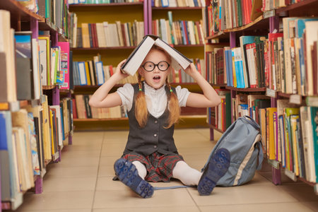 A smiling schoolgirl in uniform sits on the floor in the library and holds a book on her head. Educational concept. Modern generation of children.の写真素材