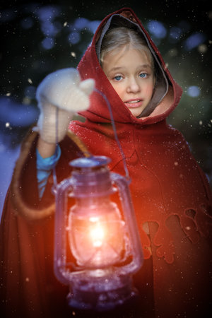Old Christmas Tale. Cute medieval girl in winter clothes with a cape and a hood walks through the snowy forest with a burning lantern. Historical reconstruction.の写真素材