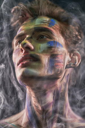 Body painting project. Portrait of a handsome man with colorful painted face looking dreamily in front of him. Art and fashion. Black background with smoke.の写真素材