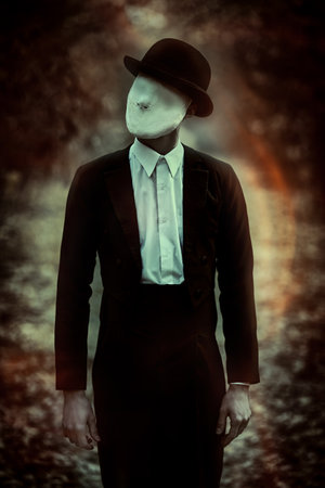 A strange and creepy gentleman in a mask of plaster bandage on his face, in a bowler hat and a black tailcoat, stands in an old autumn park. Halloween. Vintage style. Horror, thriller.の写真素材