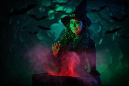 Halloween night. A beautiful witch in a black dress and hat conjures over a cauldron with a magic wand on a dark night. Magic.の写真素材
