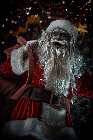 Christmas and Halloween. Scary Zombie Santa Claus is standing with a bag of gifts behind his back in a room with a Christmas decoration, entangled in cobwebs, and smiling evilly.の写真素材
