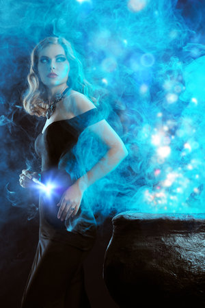 Halloween magic. A beautiful young witch in a hat and elegant black off-the-shoulder dress boils a potion in a cauldron. Art of witchcraft.の写真素材