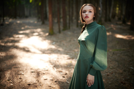 A young beautiful girl in a long green dress walks in an enchanted forest lit by sunlight. Fantasy, fairy tale heroine. Copy space.の写真素材
