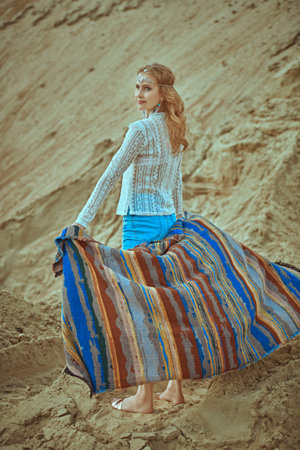 A pretty carefree girl in boho style clothes and jewelry stands in a desert among sand dunes holding an ethnic headscarf in her hands. Free lifestyle.の写真素材