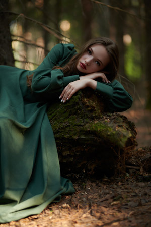 Portrait of a lovely fairy tale girl with brown hair in a long green dress lying on a large fallen tree in a dark forest. Fantasy, fairy tale heroine.の写真素材