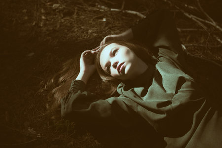 Fantasy, fairy tale heroine. An enchanted girl in a green dress is lying on the ground in a dark magical forest lit by sun beams. Legends and fairy tales.の写真素材