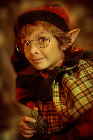A cute little elf boy stands outdoors on a snowy night cheerfully looking at the camera. Christmas...