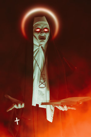 Halloween. Horrors. An evil cursed nun with red burning eyes holds a bible and a cross in her hands standing on a black background with red haze.の写真素材