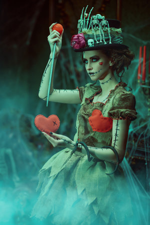 A beautiful and treacherous young woman dressed as a voodoo doll stands in an old abandoned castle and sticks a pin into the victim's toy heart. Voodoo religion. Halloween.の写真素材