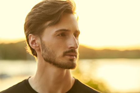 Close up portrait of a young man standing thoughtfully outdoors on the background of a river at sunset. People and emotions.の写真素材
