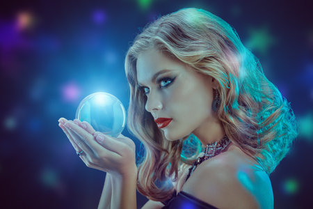 Magic time. Beautiful young woman in a black dress with elegant makeup holds a shining crystal ball. Dark blue background. Party poster. Halloween.の写真素材