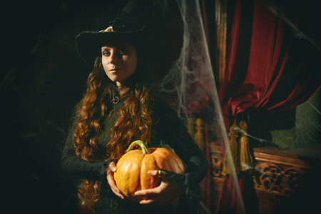 Halloween time. Pretty young witch with long curly hair in a black dress and hat holds a pumpkin...
