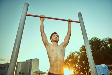 An athletic young man with bare torso is doing pull-up exercises on the horizontal bar in the park...