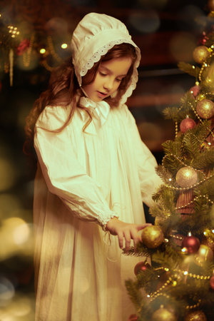 A cute little girl in a nightie and a nightcap curiously looks at the beautiful decorations hanging on the Christmas tree. Vintage style. Magic Christmas.の写真素材