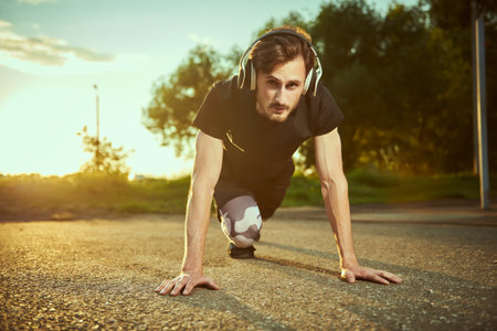 Sporty lifestyle. An active guy in sportswear performs physical exercises on the ground in the park listening to music on his headphones.の写真素材
