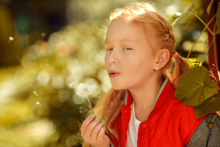 A red-haired little girl in a stylish jacket blows joyfully on a dandelion on a sunny summer day. Happy children. Carefree summertime.の写真素材