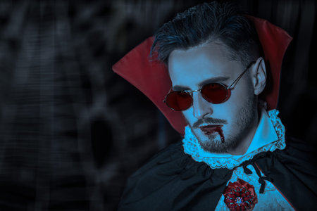 Sinister bloodthirsty dracula in an elegant suit and cloak with stylish red glasses and blood on his...