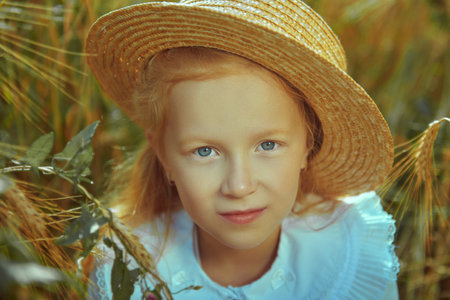Close up portrait of a lovely little girl with red hair in elegant white shirt and straw hat sitting in a wheat field. Summer day. Children's fashion, romantic style.の写真素材
