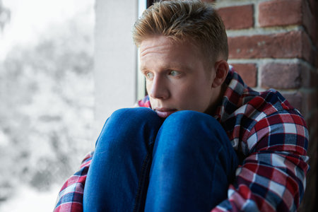 A sad guy in casual clothes sits lonely on the windowsill hugging his knees and looking thoughtfully...