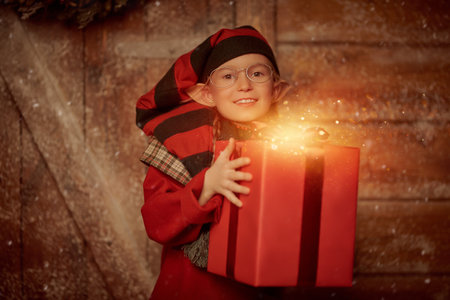 Merry Christmas. A cheerful little elf girl stands at a wooden hut in the winter forest with a red gift box. Christmas preparations and gifts.の写真素材