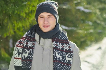 A handsome guy in a winter jacket, knitted hat and scarf stands by snow-covered fir trees in a winter park. Winter fashion.の写真素材