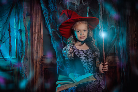 Pretty little girl in a witch costume conjures with a book of spells and magic wand in a dark old house covered in cobweb. Halloween time.の写真素材