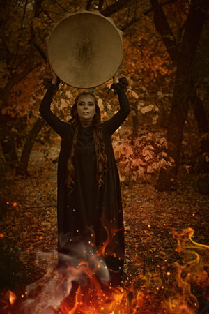 A female shaman in black garment with ethnic ornaments on her face performs a ritual with a shamanic drum in an autumn forest in front of a burning fire. Ethnic culture. Shamanic rituals.の写真素材