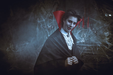 Halloween. An attractive young vampire in an elegant tailcoat and a cloak stands in an old castle...