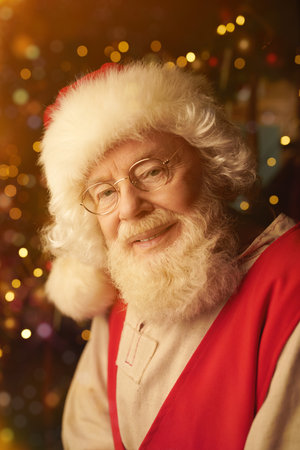 A portrait of a good old Santa Claus in a canvas shirt, a red vest and a hat, looking at the camera and smiling surrounded by shining golden lights at cozy home. Christmas legends and fairy tales.の写真素材