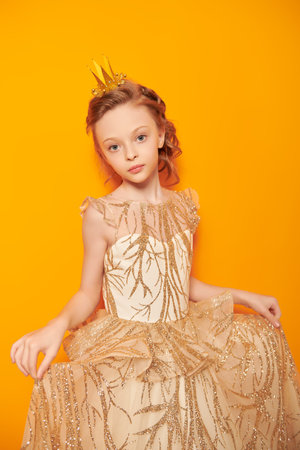 A charming little princess poses in a golden dress and a crown in a bright yellow background. Dreams and kid's fashion.の写真素材