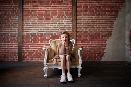 Pretty little girl dressed in elegant casual clothes sits thoughtfully in an armchair. Loft style room. Copy space.の写真素材