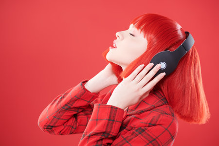 DJ, party girl. A stylish girl with bright red lips and red hair wearing a red checkered jacket enjoys music on headphones. Red studio background. Beauty, Fashion. Copy space.の写真素材