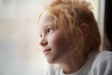 Close-up shot of a lovely red-haired little girl in white sweater looking out the window with a smile. Natural lighting. Children, childhood, emotions.の写真素材