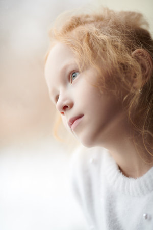 A dreamy red-haired little girl in white sweater looking pensively out the window. Natural lighting. Children, childhood, emotions. Close-up shot.の写真素材