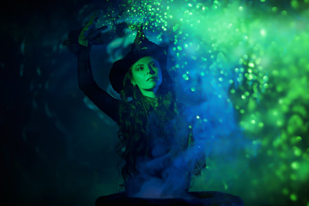 Magic on Halloween night. A beautiful witch in a black dress and hat conjures over a cauldron on a dark moonlit night.の写真素材