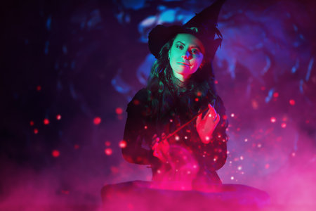 Halloween night. A beautiful smiling witch in a black dress and hat conjures over a cauldron with a magic wand on a dark moonlit night. Magic time. Art of witchcraft. Copy space.の写真素材