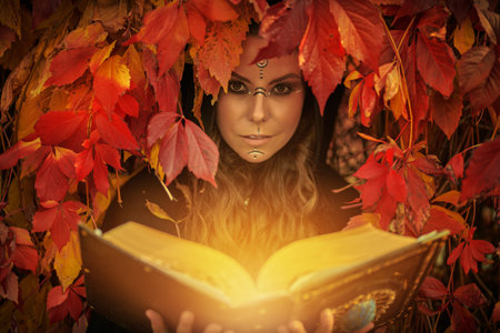 A beautiful shaman woman with long curly hair and ethnic ornaments on her face dressed in black garment stands among red foliage in the forest with a magic book. Fantasy. Ethnic culture. Shaman woman.の写真素材