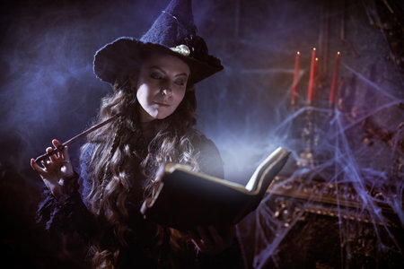 Magic Halloween spells. Beautiful curly-haired sorceress in a black hat makes a spell with a magical book and a wand in a dark old castle covered in cobweb. Art of witchcraft.の写真素材
