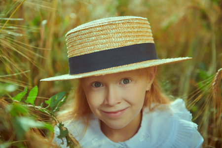 Portrait of a lovely smiling little girl with red hair in elegant white shirt and straw hat sitting in a wheat field. Summer day. Children's fashion, romantic style.の写真素材