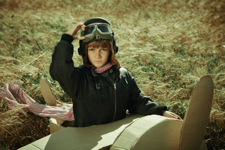A kid in a leather jacket and aviator's helmet playing in a field with a cardboard plane. Children's...