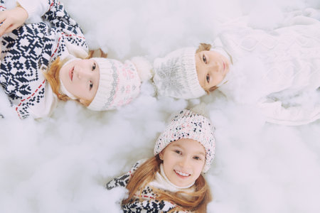 Children's winter fashion. Three cute, joyful children in knitted sweaters and hats lie together on the snow and smile. Studio portrait. Winter activities.の写真素材