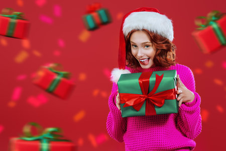 A cheerful pretty girl with bright red hair is holding a gift and sticking out her tongue for joy. Festive red background with copy space and a lot of gift boxes around. Christmas sales and shopping.の写真素材