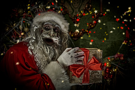 Christmas and Halloween. Scary Zombie Santa Claus is standing with a gift box in a room with a Christmas decoration, entangled in cobwebs, and smiling evilly.の写真素材