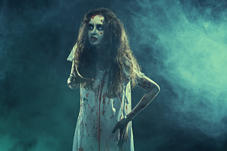 Creepy zombie woman. A terrible disfigured woman with faded eyes, possessed by the devil, in a bloody nightie bends, standing in the dark, surrounded by smoke. Horror, thriller. Halloween.の写真素材