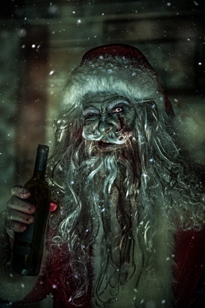 Christmas, New Year and Halloween. Merry and scary zombie Santa Claus with a bloody beard stands by his wooden hut with a bottle of alcohol and laughs wickedly.の写真素材