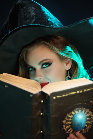 Attractive young sorceress in a black hat looking from behind a Book of Spells. Dark background with...